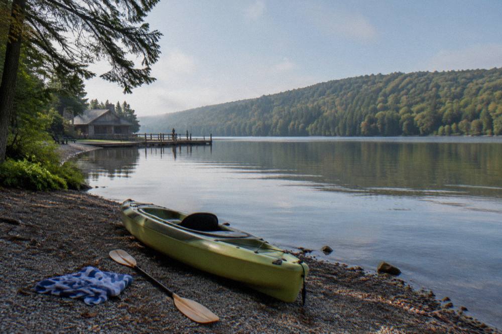 A single kayak resting on a gravel lake shore beside still water and tree-covered hills