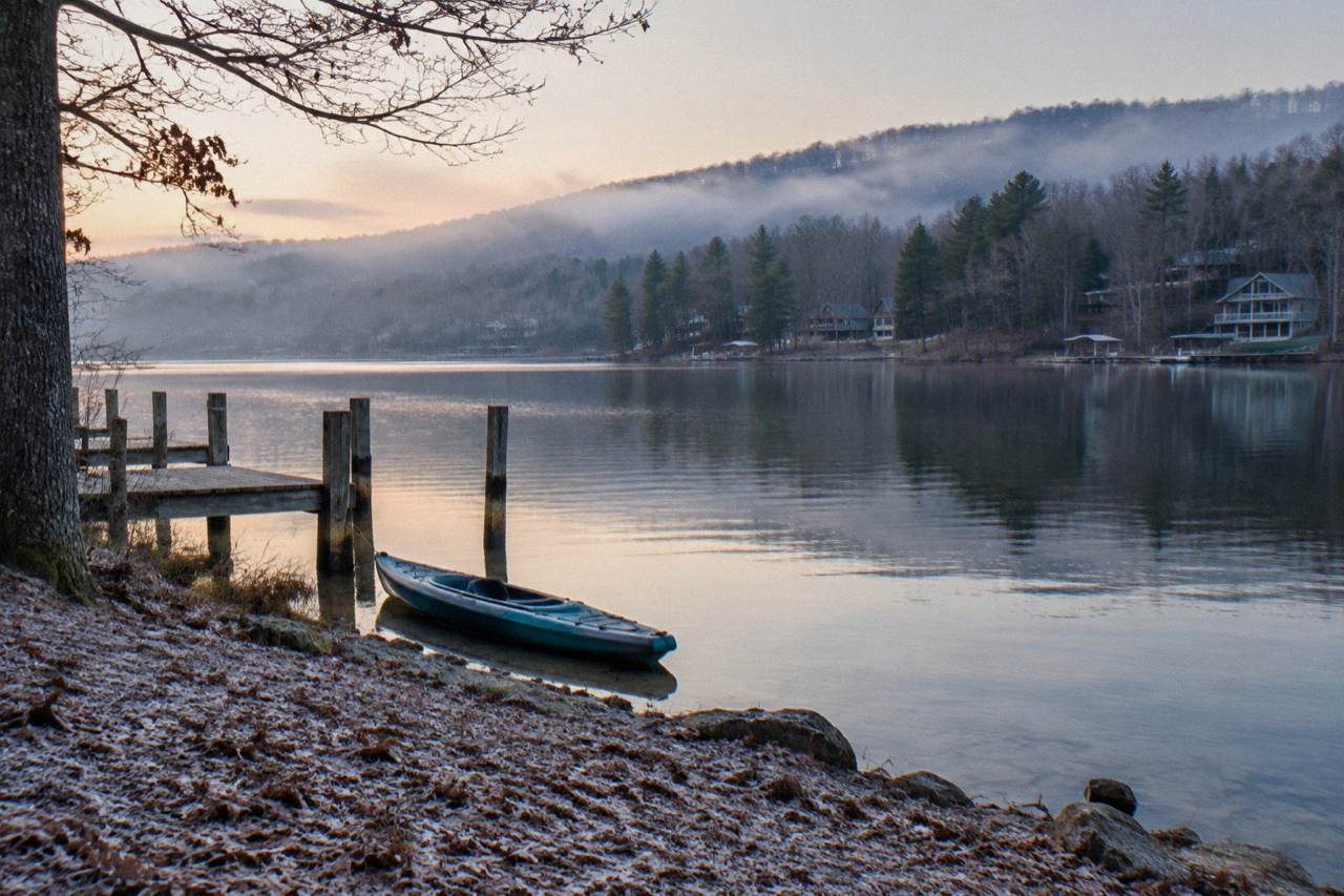 A single kayak resting on a quiet lake cove at dawn with forested hills behind it