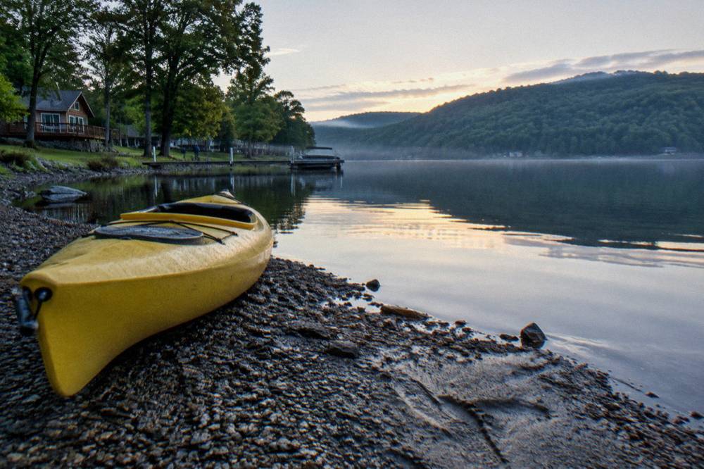 A kayak resting on a gravel shoreline beside still lake water at dawn