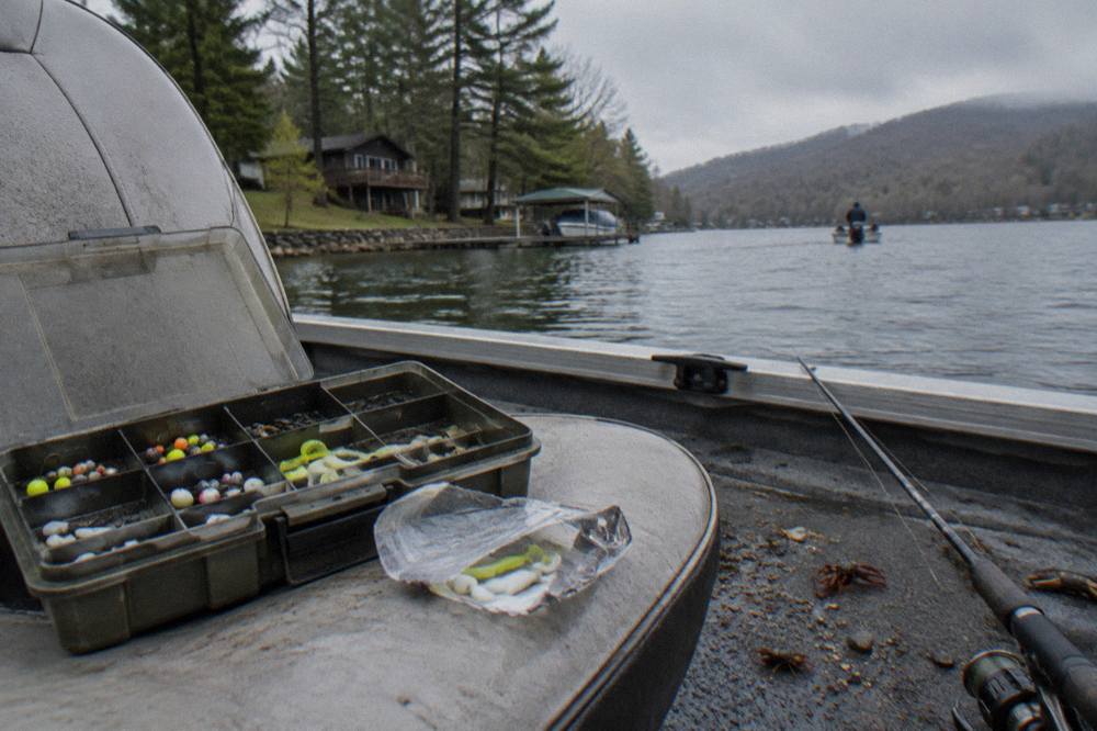 A tackle box open on a boat seat with jigs, spinners and a package of soft plastic grubs in morning light