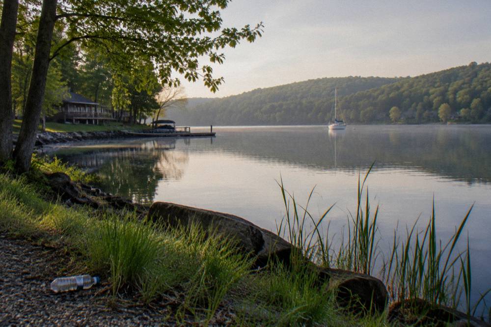 A quiet tree-lined cove with glassy water and a sailboat at anchor in the distance