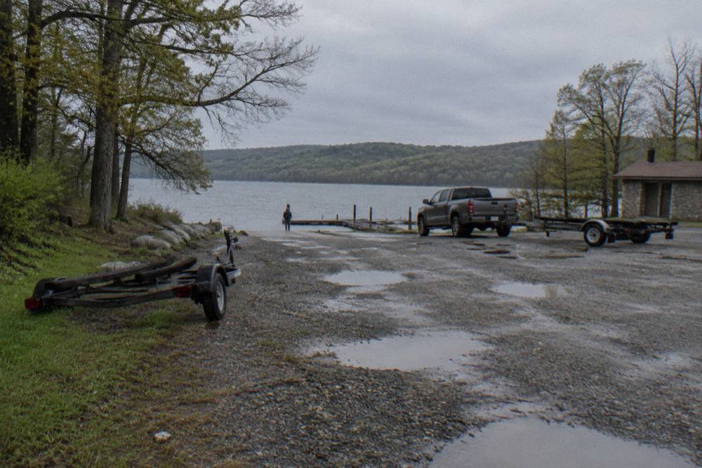 Wide view of a public boat ramp parking area with pickup trucks and boat trailers near a wooded shoreline