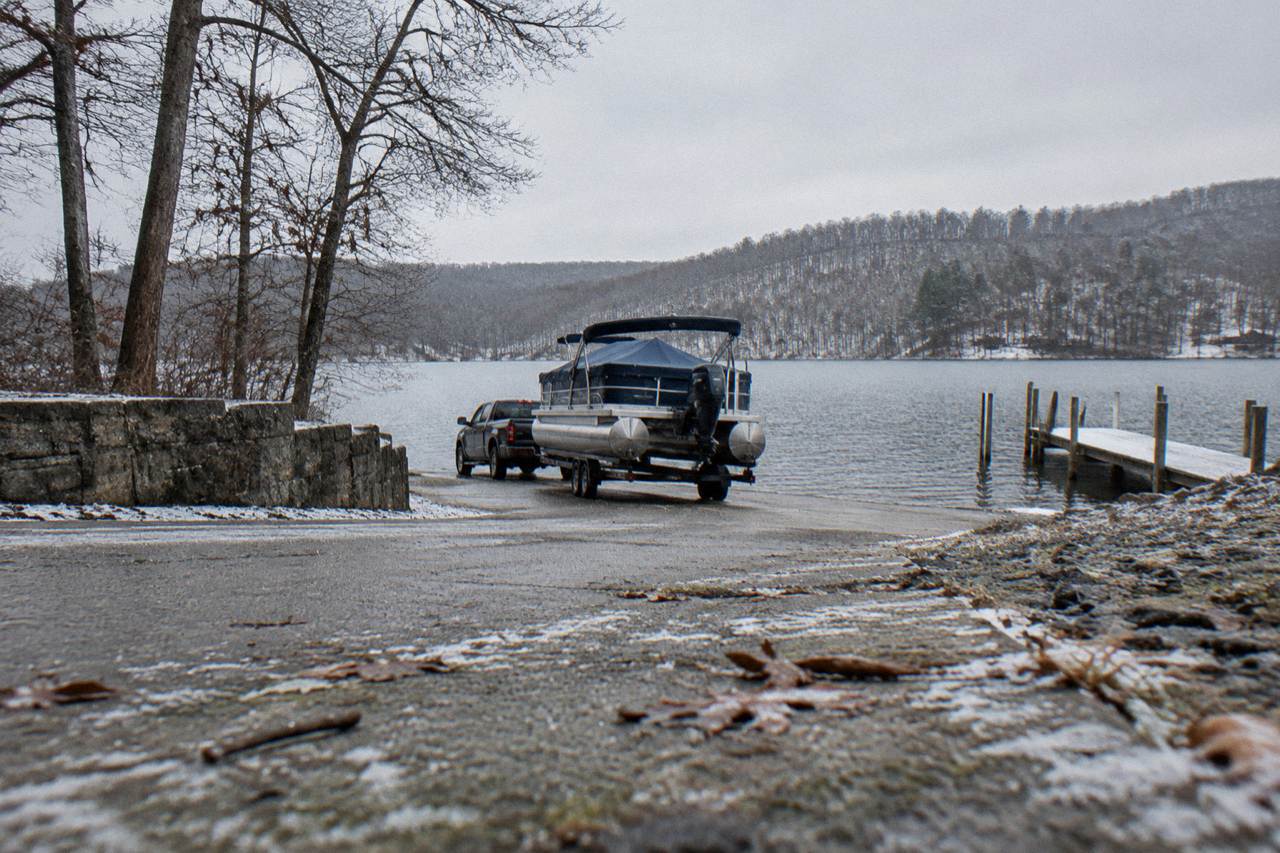 Concrete boat ramp at a wooded lake shore with a trailer backing a pontoon boat toward calm water