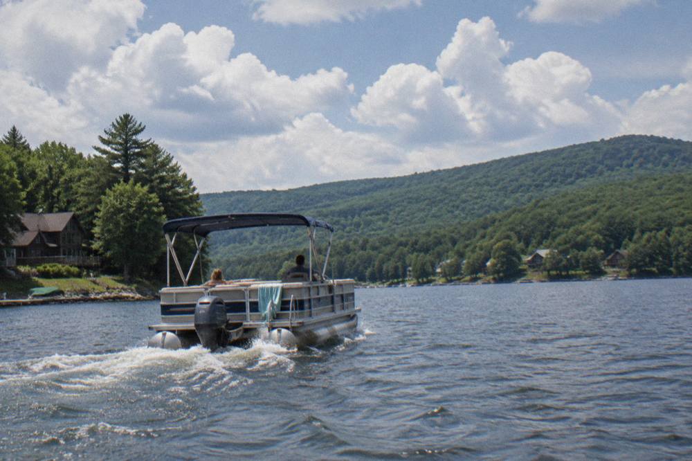 A pontoon boat cruising across open water with forested hills behind it