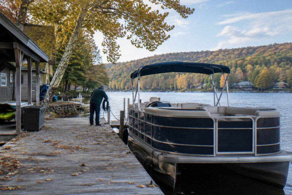 A pontoon boat tied at a wooden dock under trees turning yellow in autumn