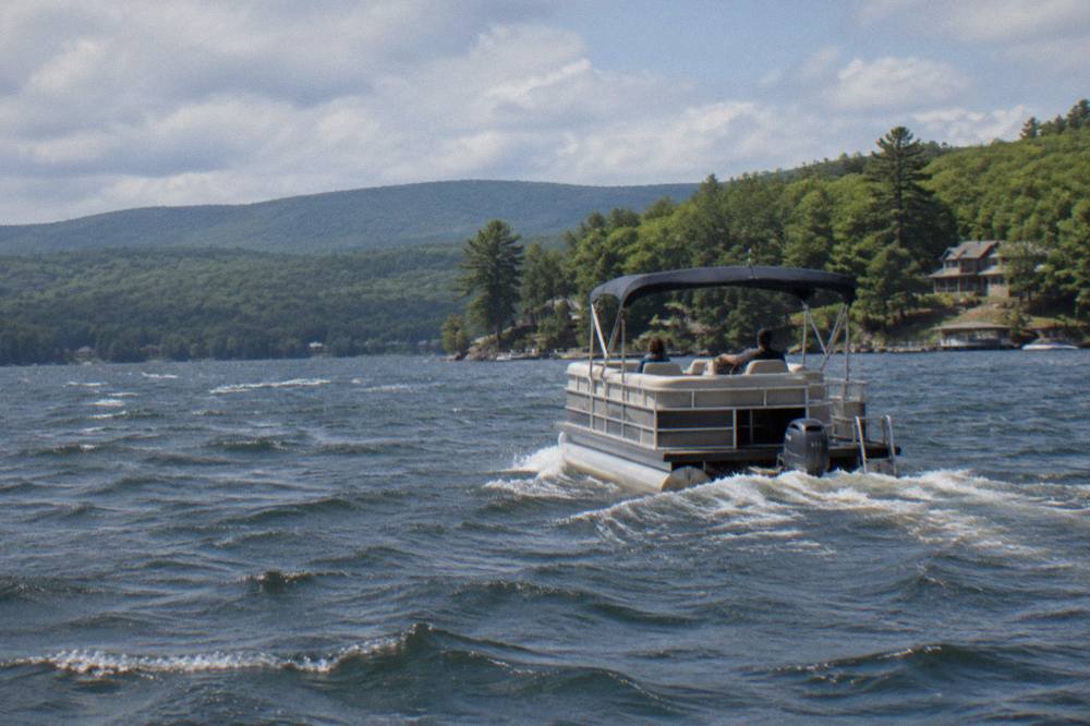 A wide pontoon boat gliding across open water with forested ridges in the background