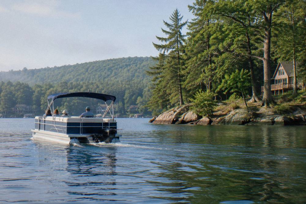 A guided pontoon boat with a canopy passing a rocky point on a calm lake morning