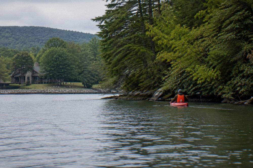 A paddler in a life jacket moving a kayak across quiet water near a wooded bank