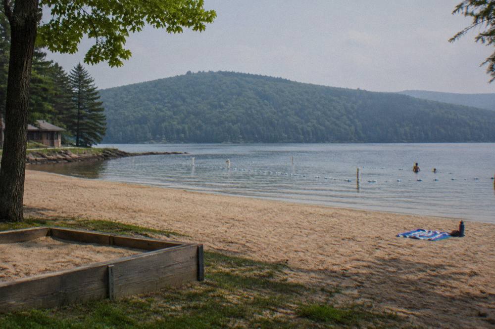 Wide view of a sandy swim beach with calm water and tree covered hills beyond